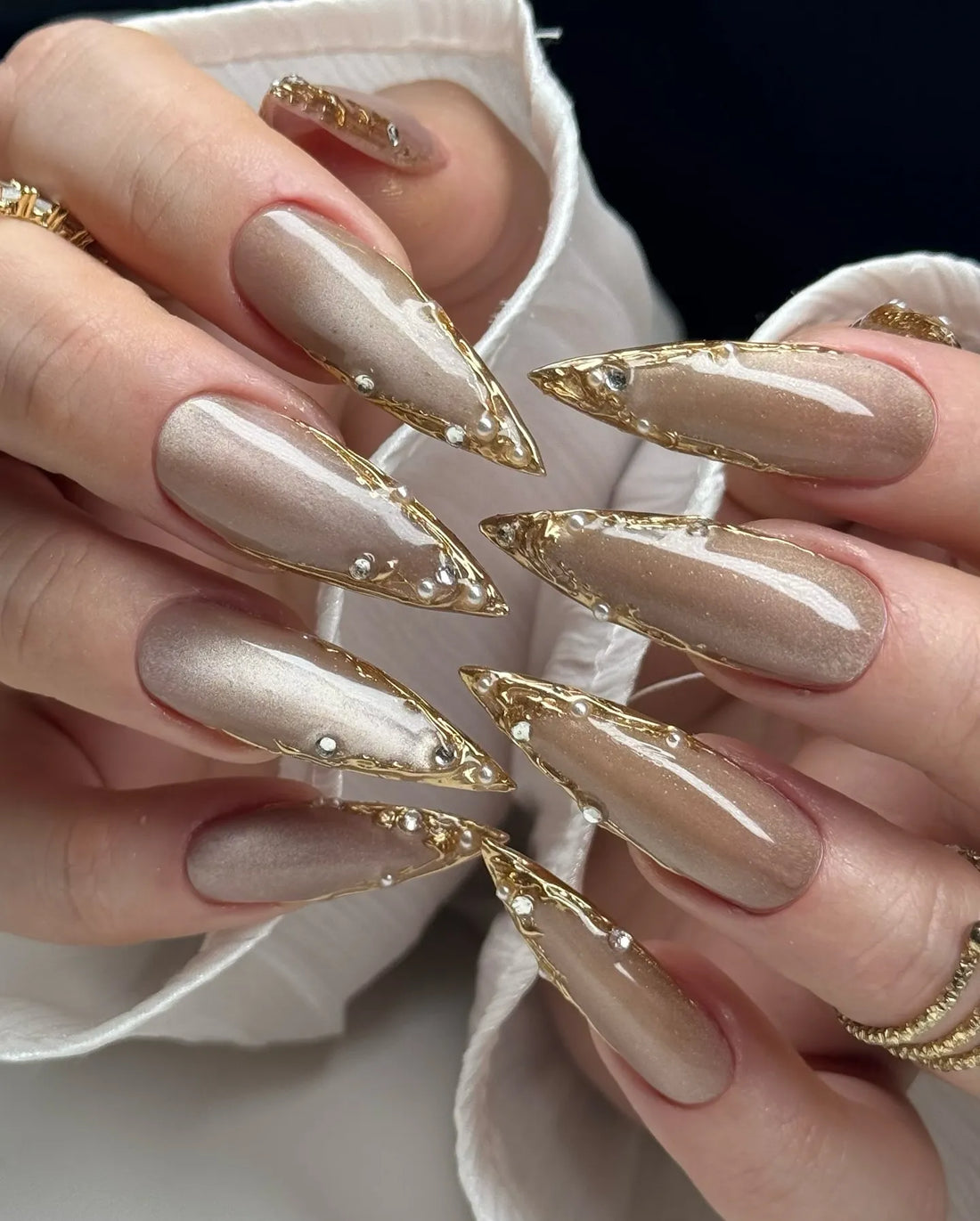 Hand with long stiletto press-on nails in metallic taupe, gold chrome tips, rhinestone accents.