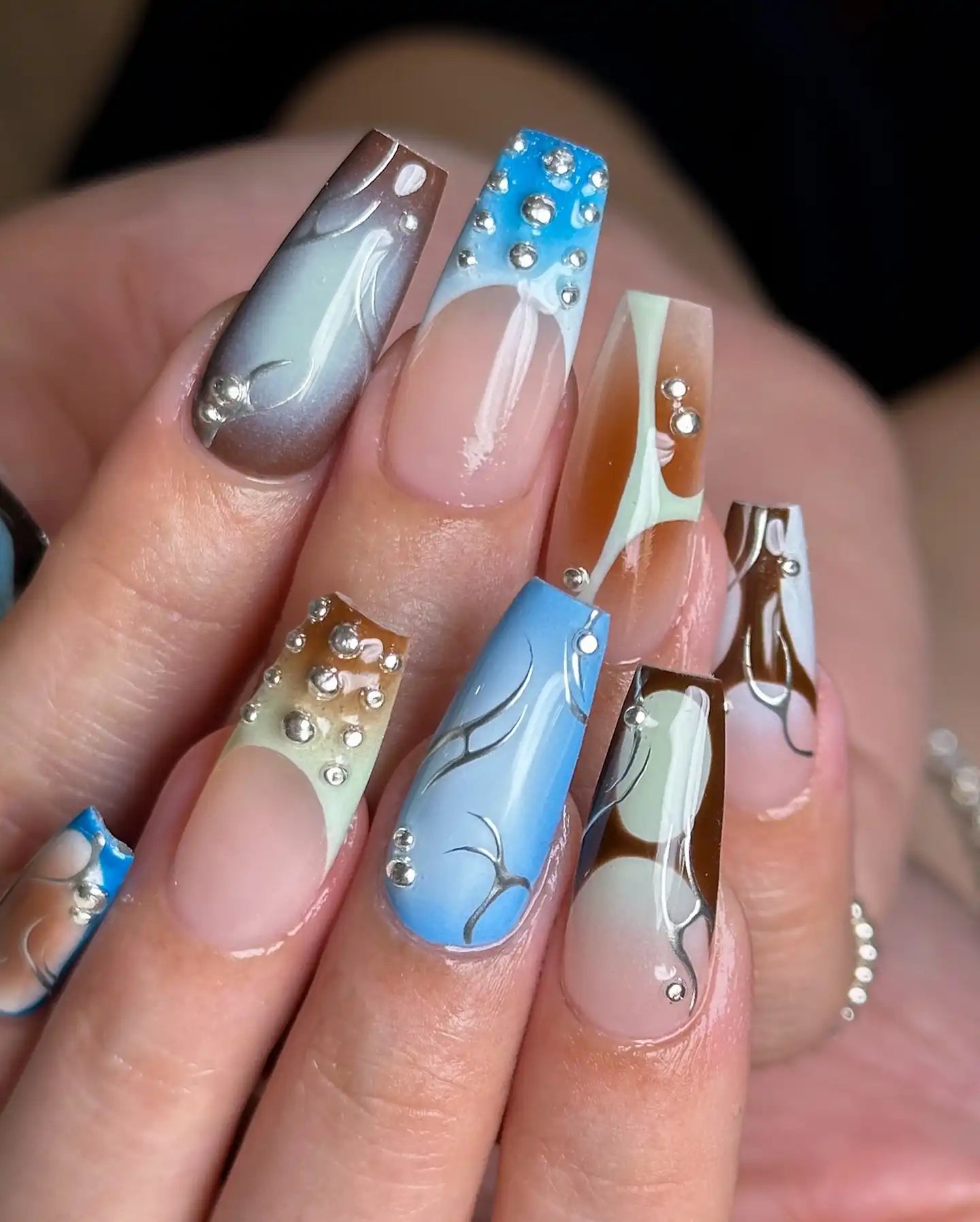 Hand with long press-on nails featuring blue, brown, and ombre designs, silver accents, and rhinestones.