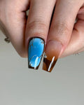 Hand with square-shaped ombre press-on nails, blue and brown designs, silver accents, rhinestones