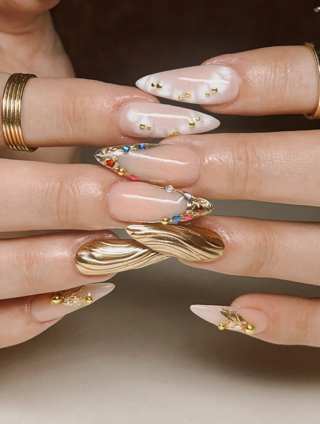 Hand with long stiletto press-on nails featuring gold chrome, marble white, and multicolor gemstones.