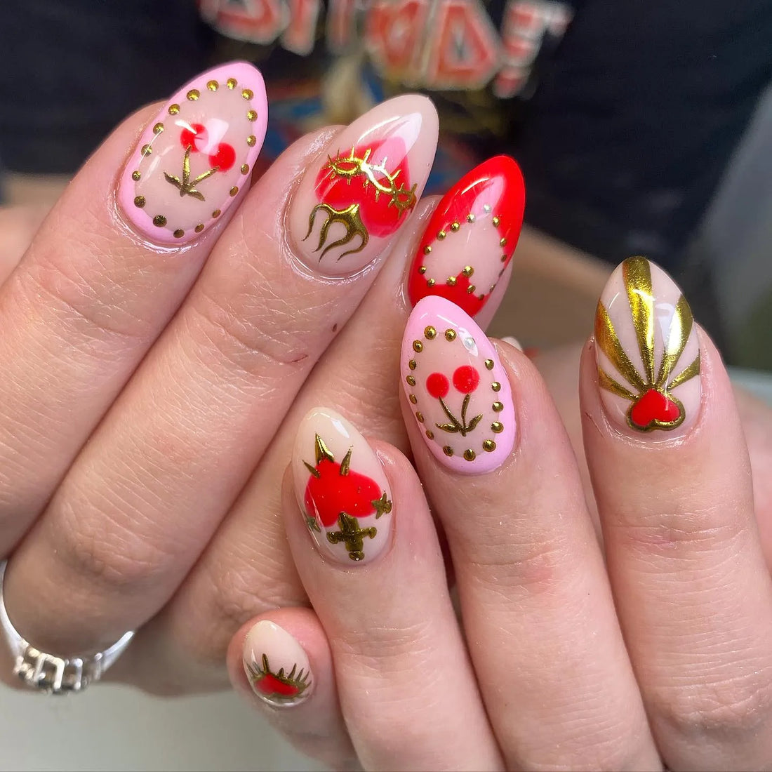 Handmade press-on nails with red hearts, gold dagger, cherry, and gothic designs on nude base.