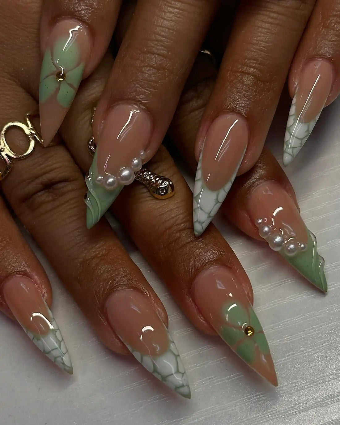 Hand with stiletto press-on nails featuring green floral accents, pearls, and crackle design tips.