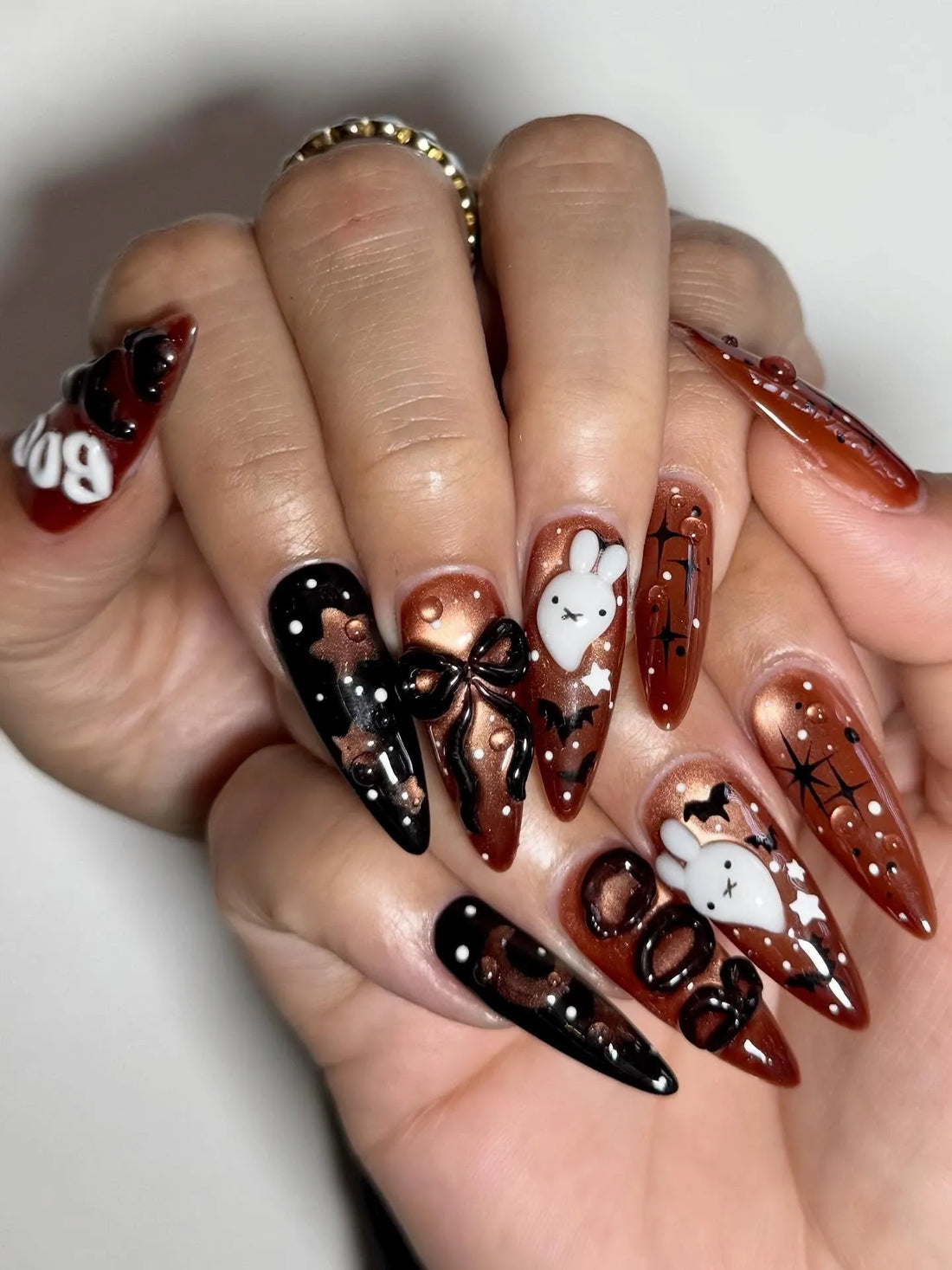 Hand with long stiletto press-on nails in black and bronze, decorated with 3D Halloween ghost, bow, bats, and spiderweb designs.