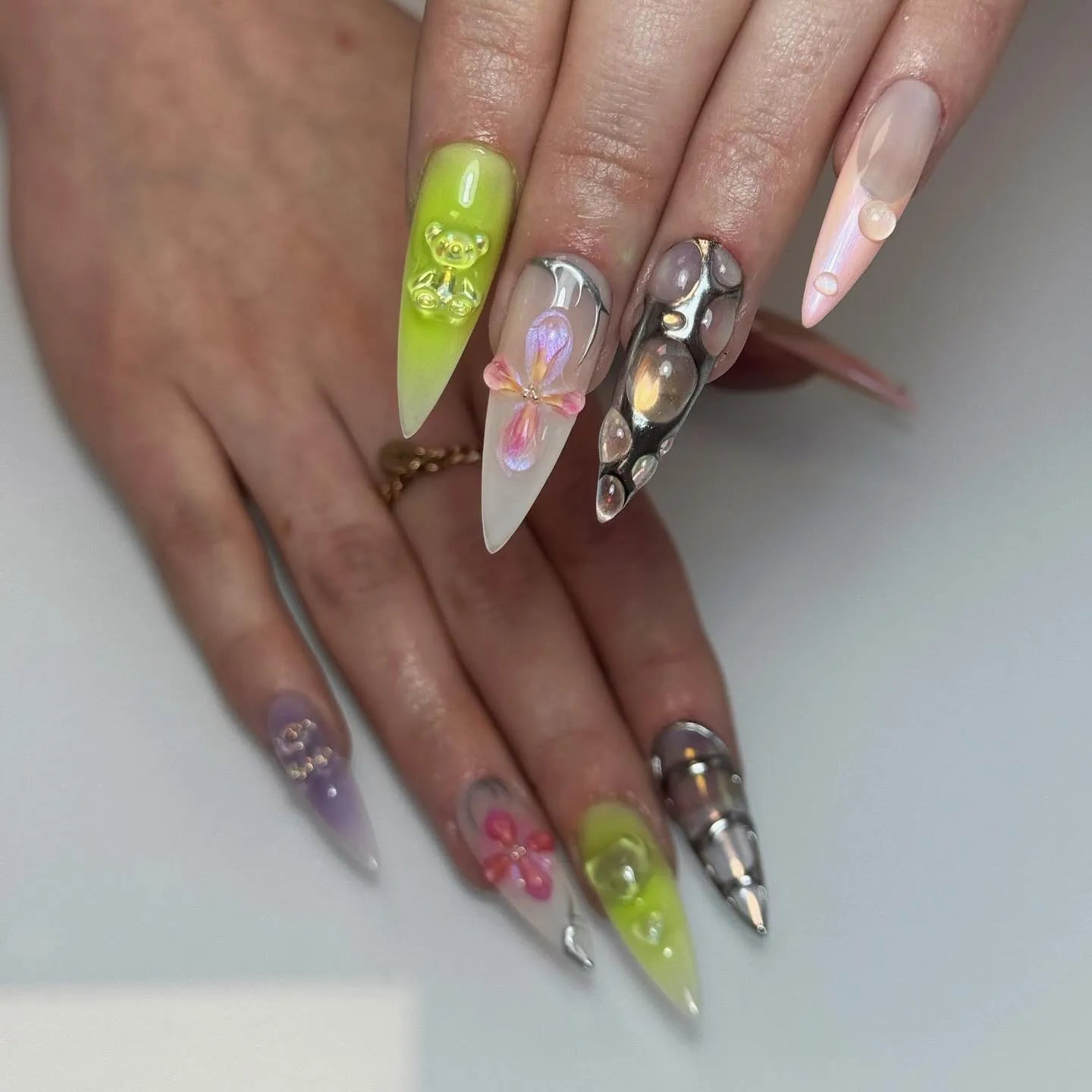 Hand with long, stiletto-shaped press-on nails featuring iridescent 3D bear, flowers, silver metallic, and cyber-inspired designs.