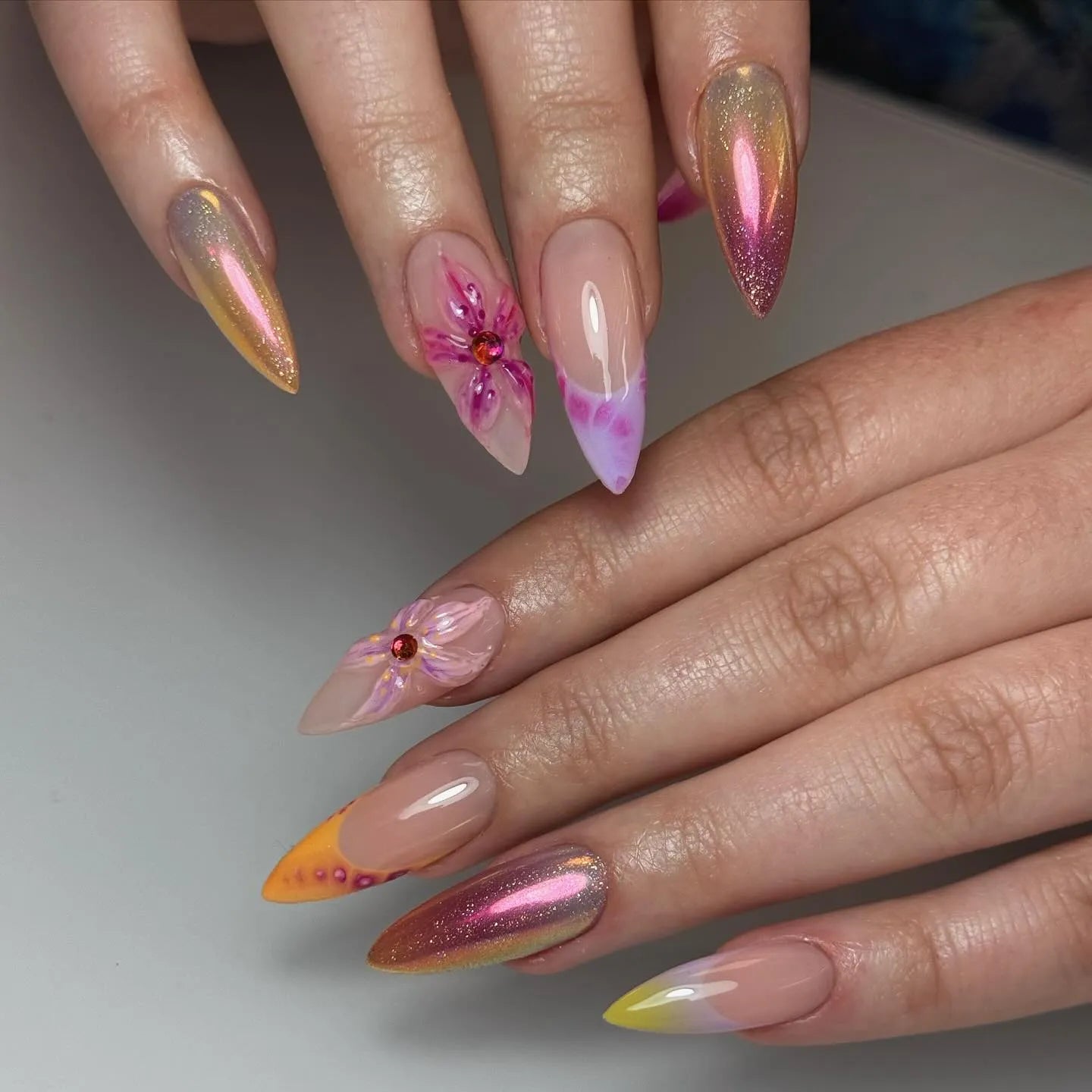 Hand with iridescent pink chrome press-on nails, 3D floral accents, and sunset ombre tips