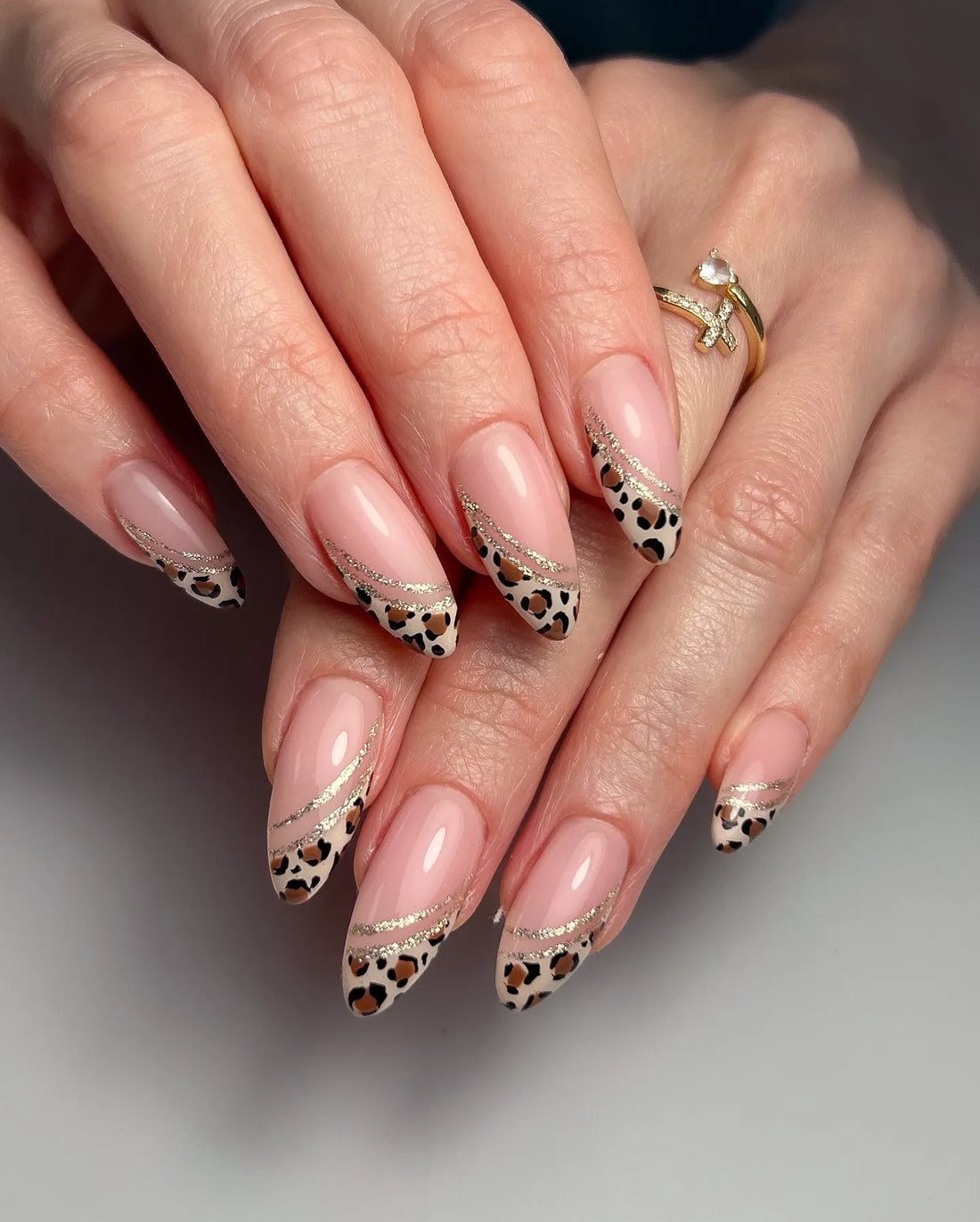 Hand with almond-shaped press-on nails featuring leopard print French tips and gold glitter swirls.