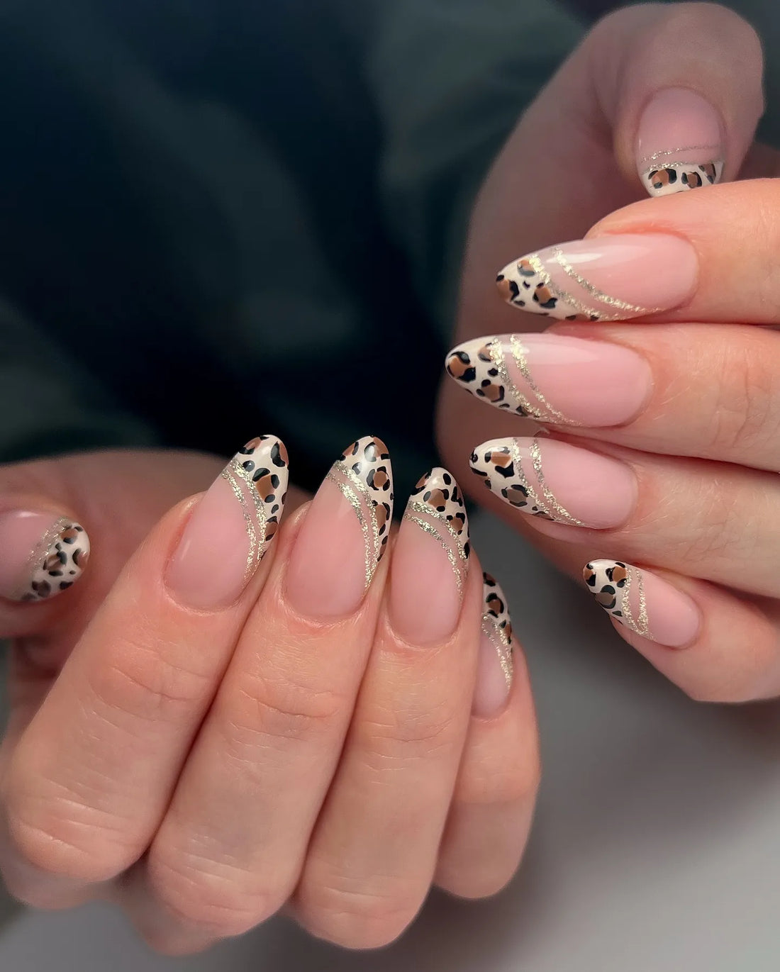 Hand with almond-shaped press-on nails featuring leopard print French tips and gold glitter swirls.