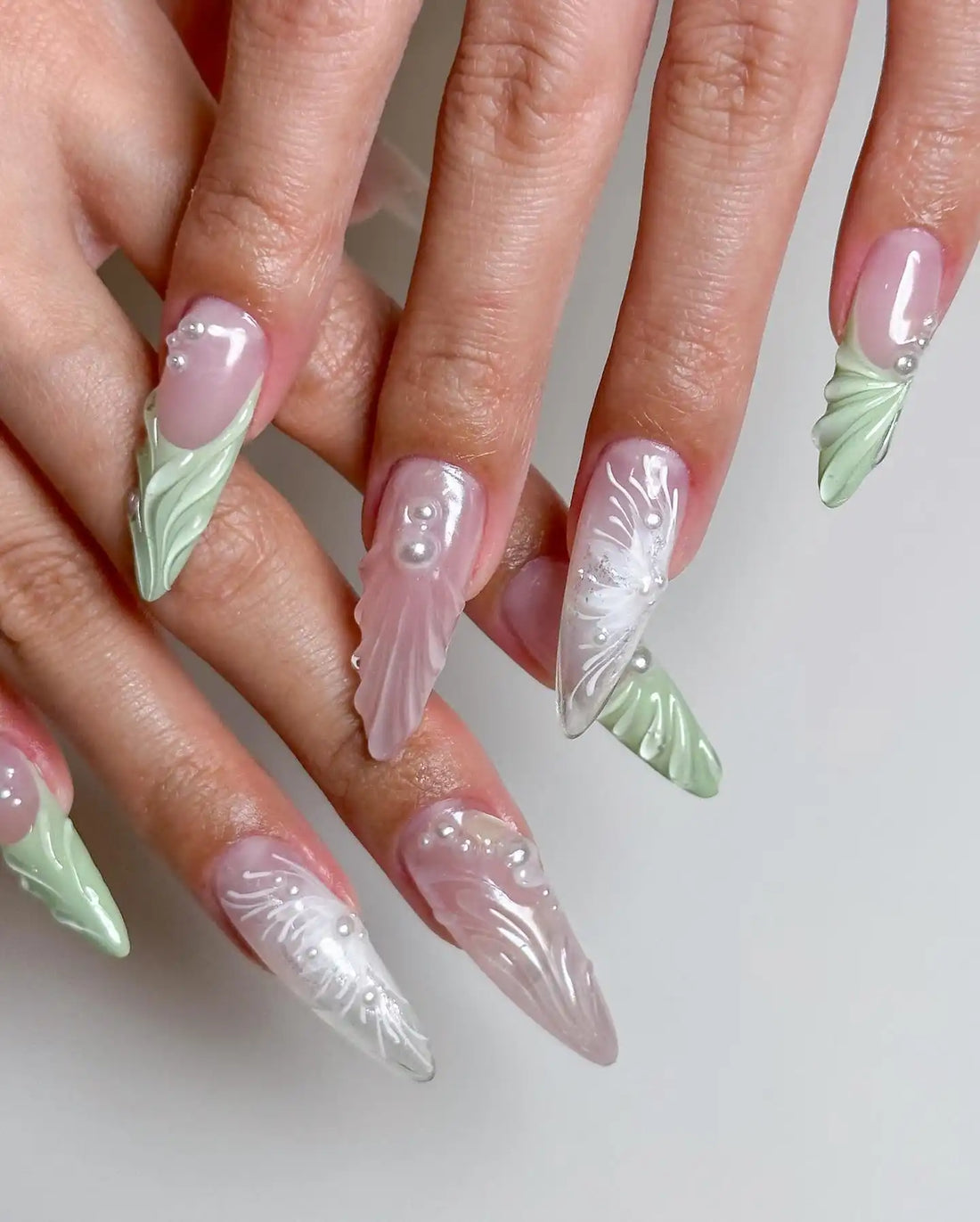 Hand with long almond-shaped press-on nails in pastel pink and green, decorated with pearls and 3D mermaid designs.