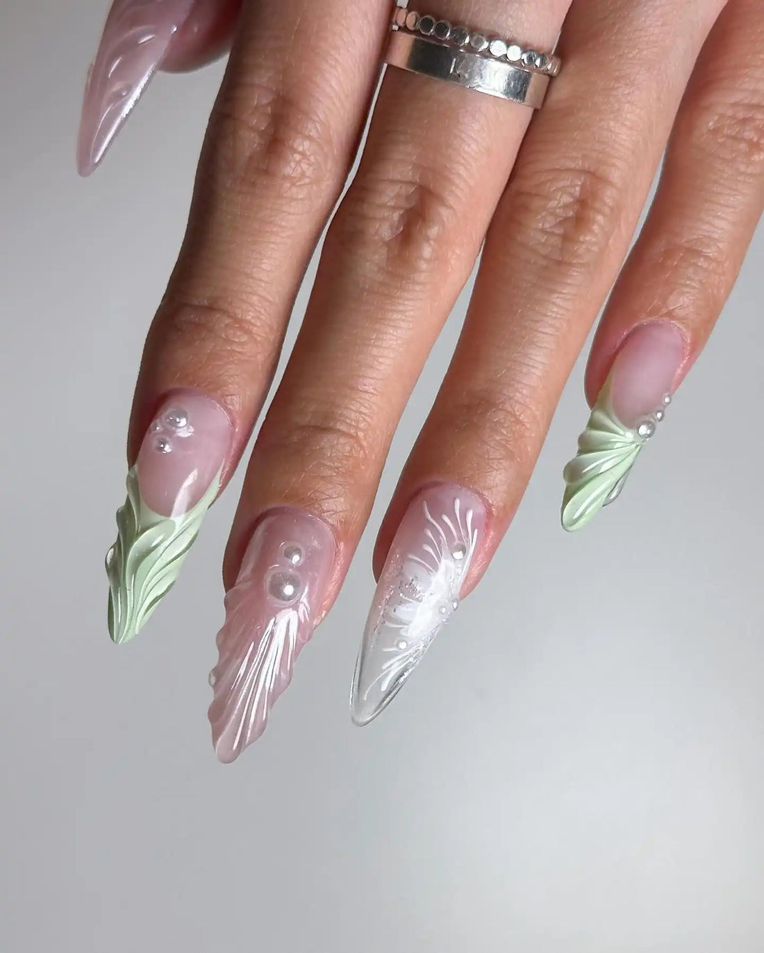 Hand with long almond-shaped press-on nails in pastel pink and green, decorated with pearls and 3D mermaid designs.