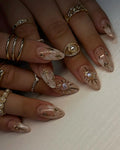 Handmade nude press-on nails with gold celestial heart and star designs, surrounded by rings.