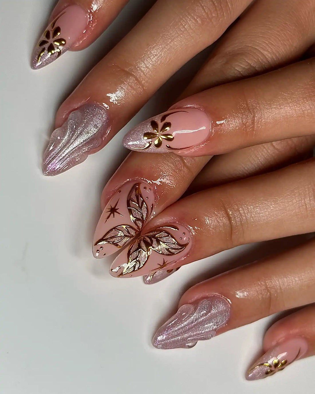 Hand with long pink nude press-on nails featuring gold butterfly and floral designs, chrome shimmer accents.