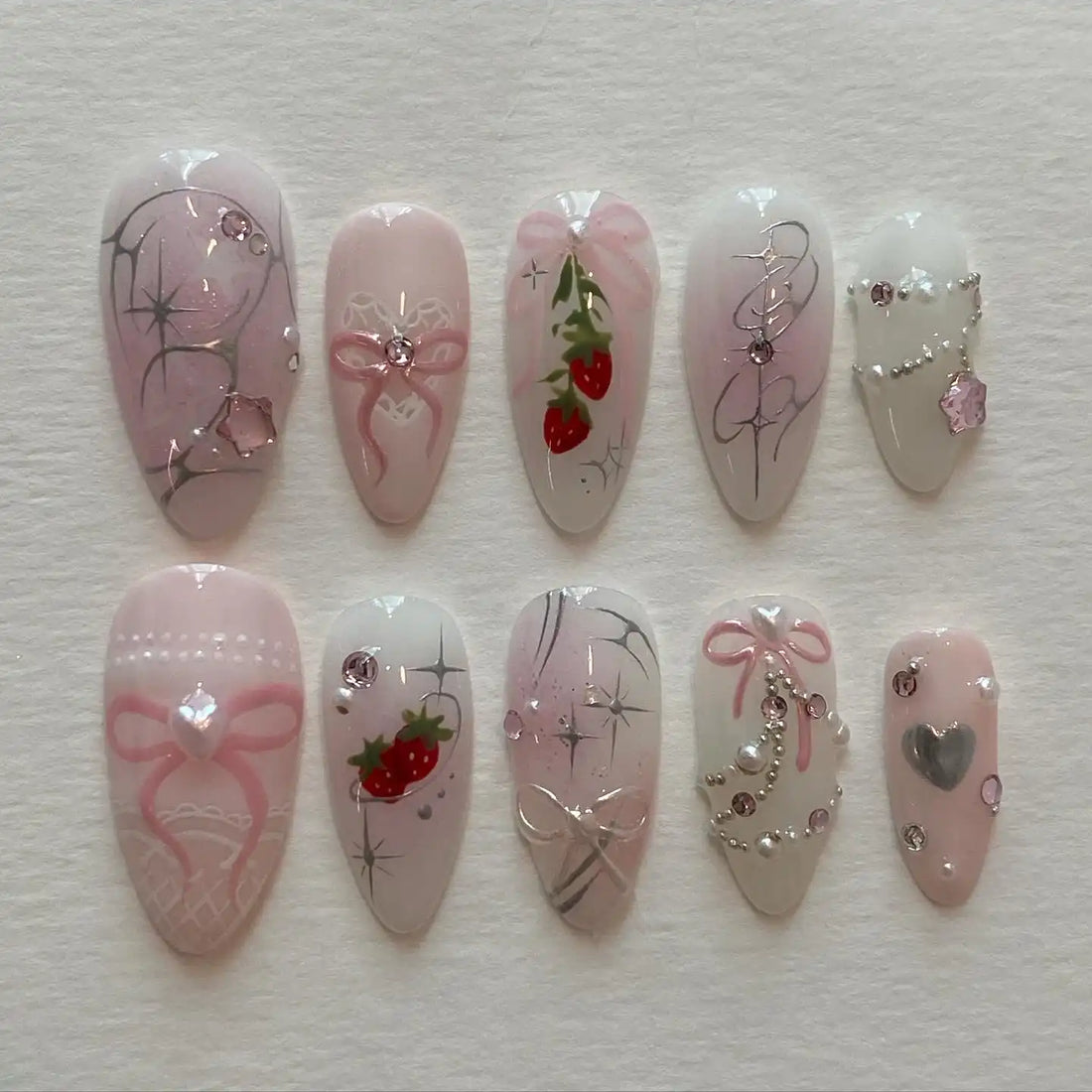 Handmade pink press-on nails with strawberry, bow, crystal, and pearl designs on white background.