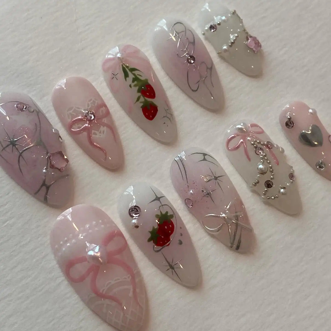 Handmade pink press-on nails with strawberry, bow, crystal, and pearl designs on white background.