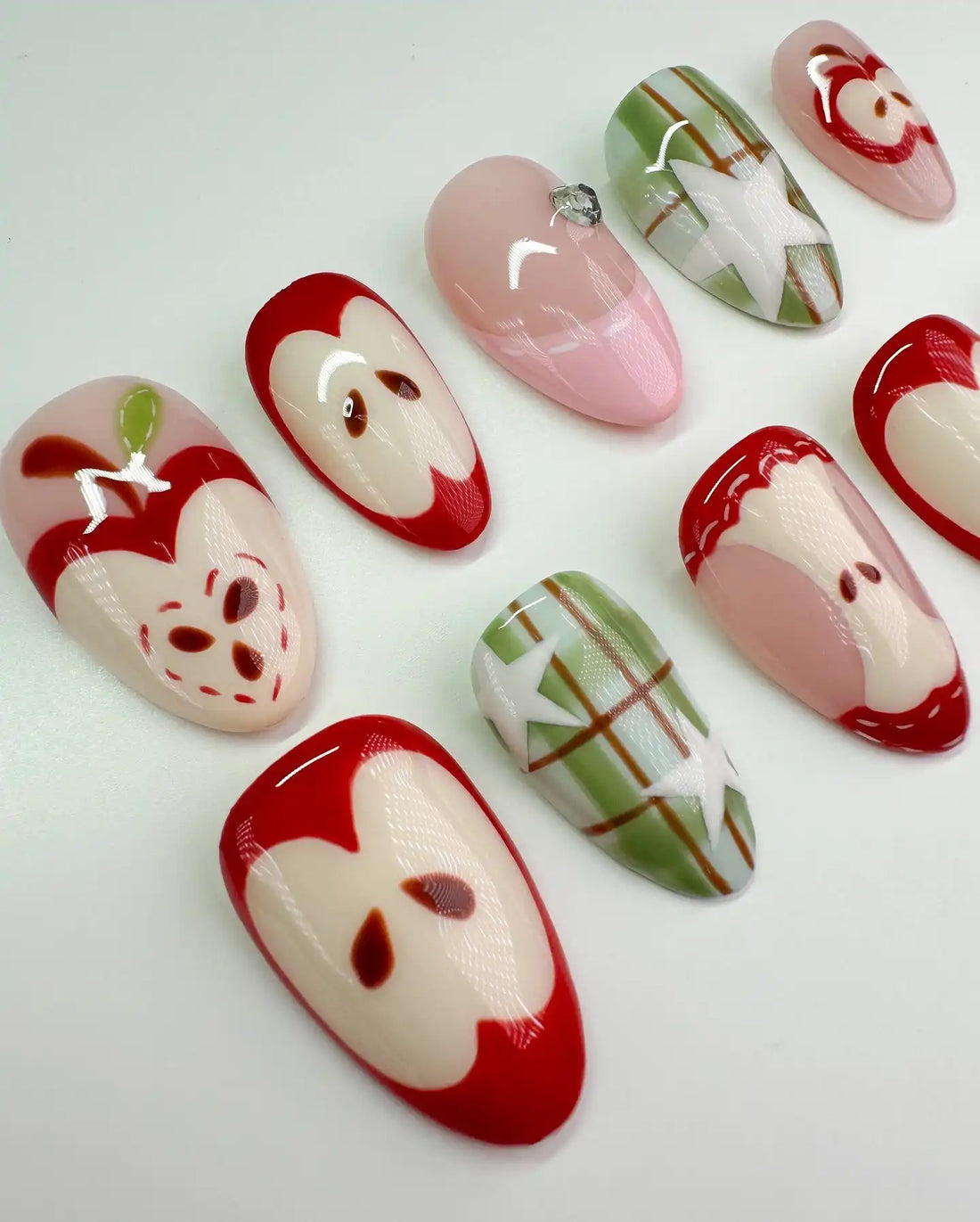 Handmade press-on nails with red apple, plaid green, and star designs, arranged on white background.