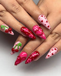 Hand with long press-on nails, pink and red heart, cherry, bow, and floral 3D nail art designs.