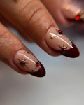 Hand with press-on nails featuring red gems, gold star designs, and wine red tips.