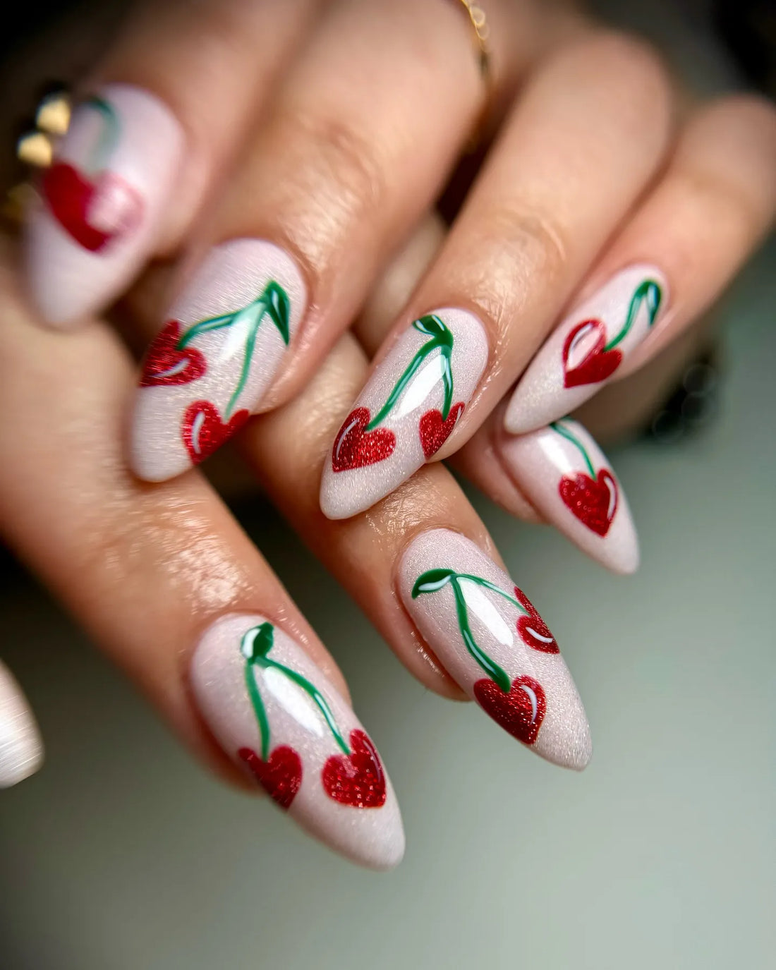 Hand with almond-shaped press-on nails featuring red glitter heart cherry designs on a white shimmer base