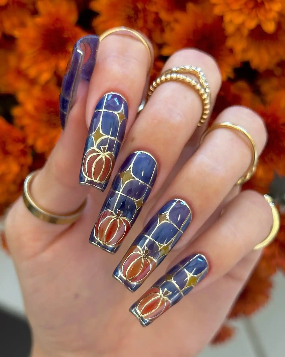 Hand with long press-on nails featuring stained glass pumpkin nail art in gold and orange, autumn flowers background.