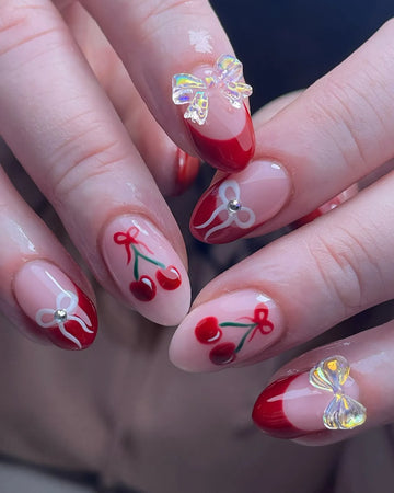 Handmade almond press-on nails with red French tips, cherry art, and iridescent ribbon bows.