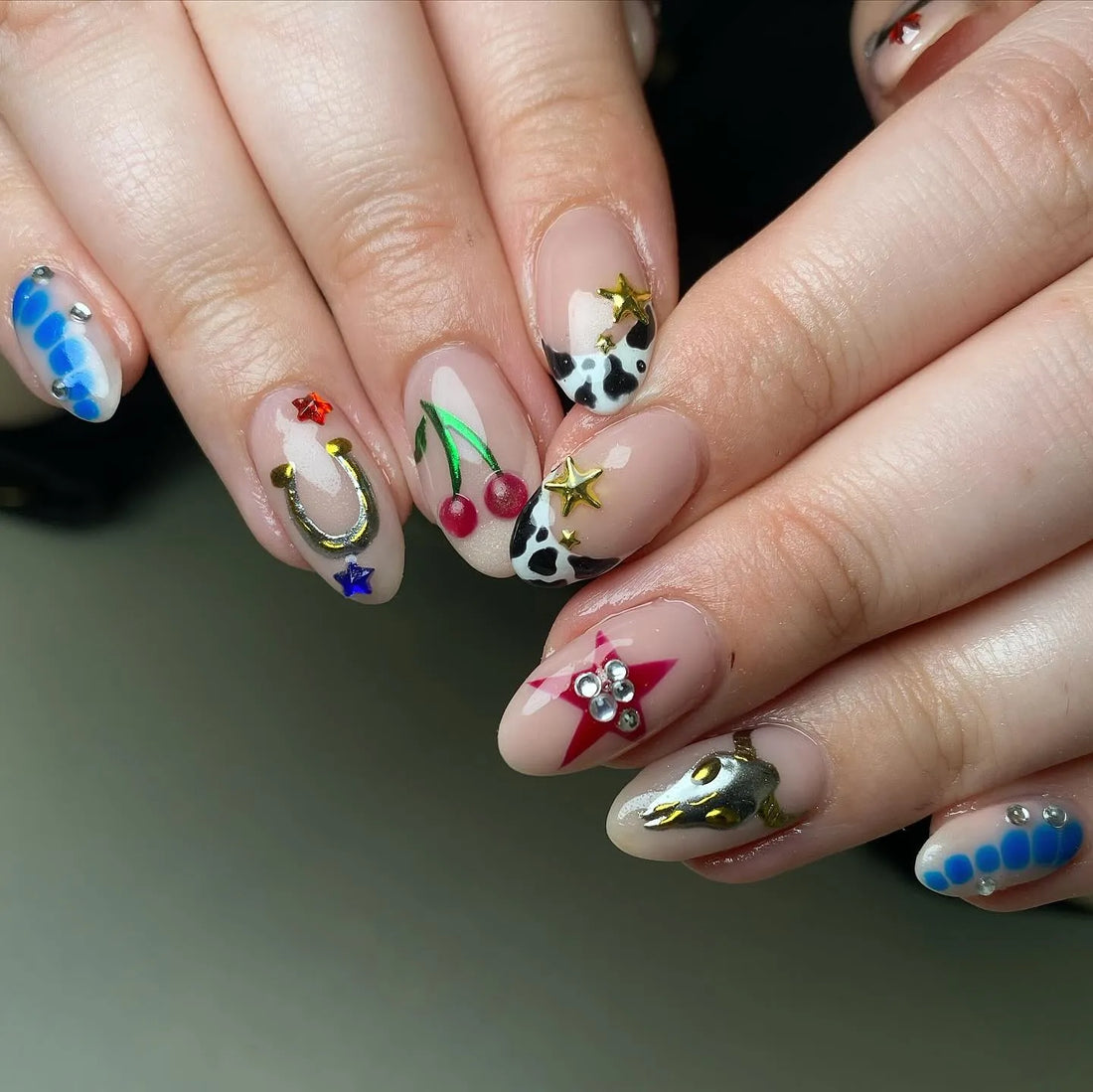 Hand with handmade western cowgirl press-on nails featuring cow print, cherries, horseshoe, and star designs.