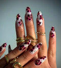 White press-on nails with pink floral design, black dots, and gold accents, worn with gold rings.