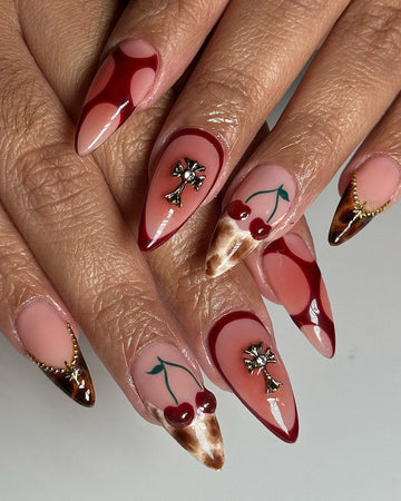 Stiletto press-on nails with cherry, cross, and gold embellishments on nude and red tips.