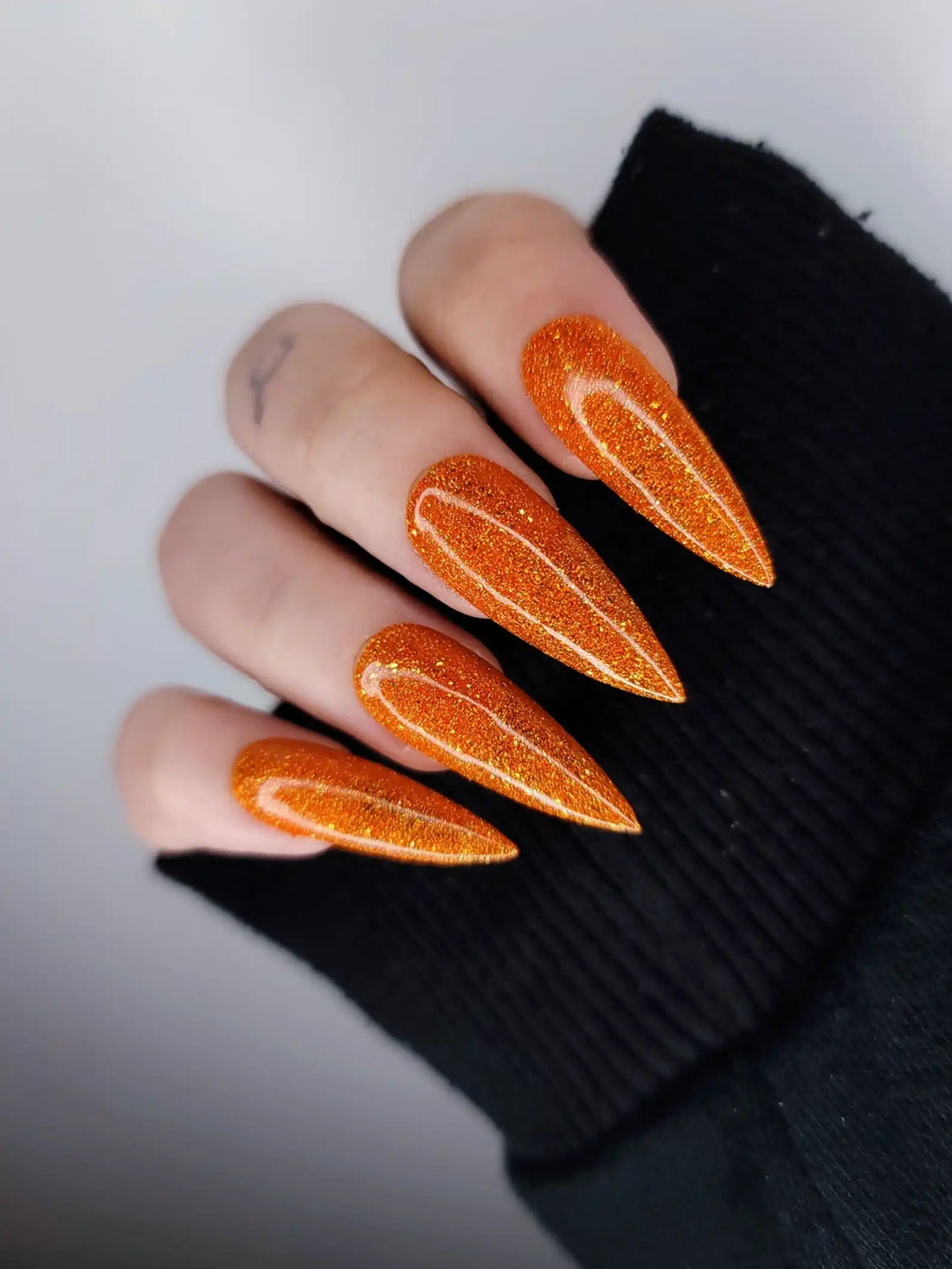 Hand with long stiletto-shaped orange glitter press-on nails, displayed on black sleeve.