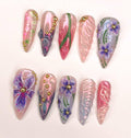 Handmade press-on nails with pink, purple floral designs, gold studs, and abstract patterns