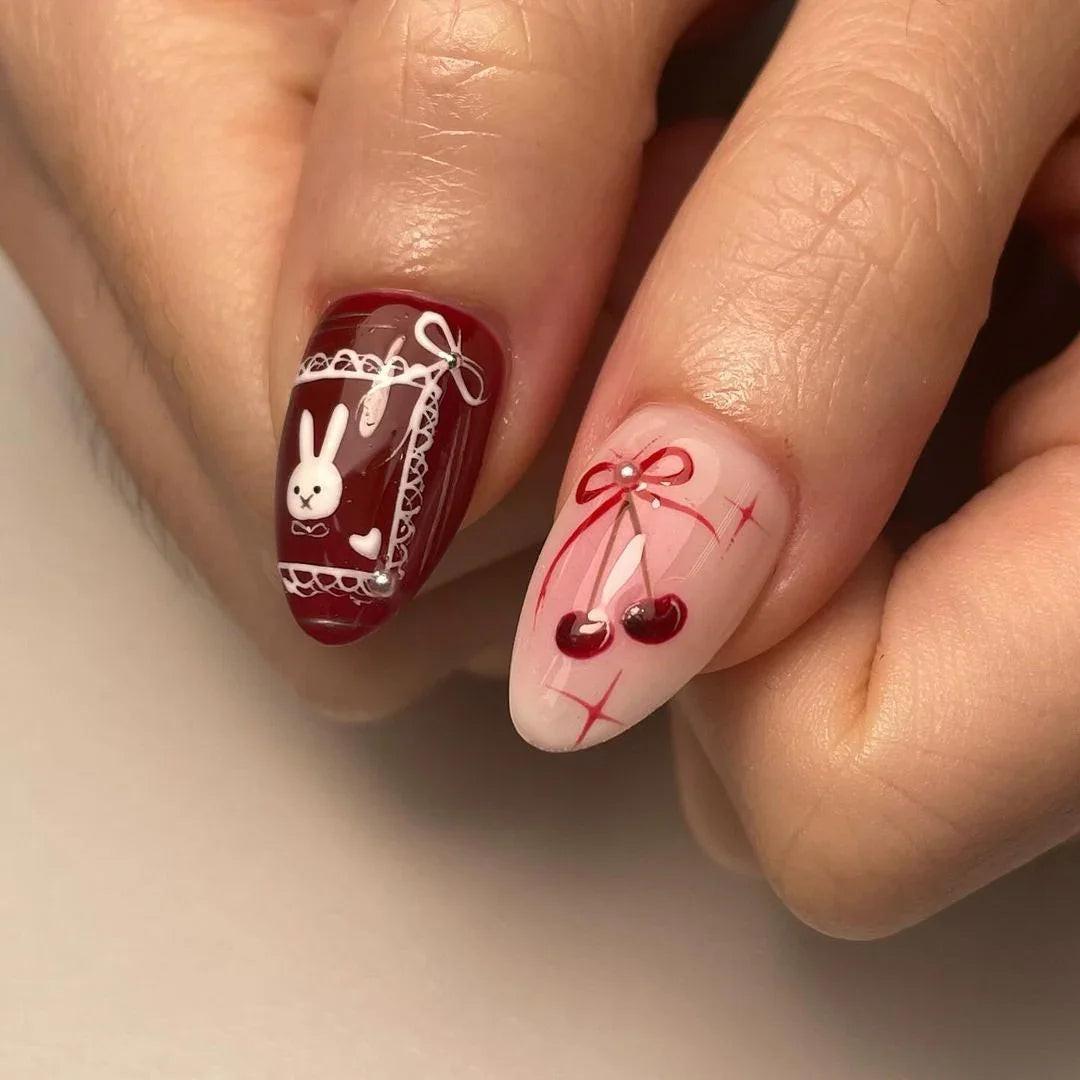 Hand with almond-shaped press-on nails in red and nude tones, featuring heart, bow, cherry, and bunny nail art designs.